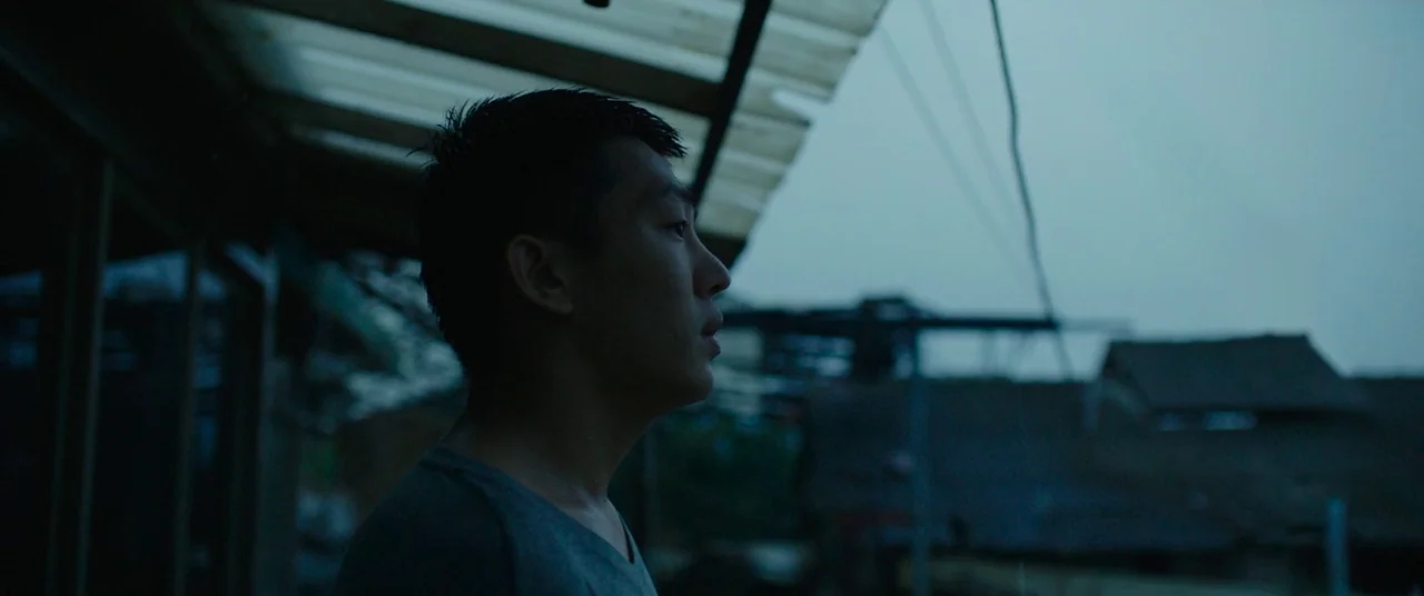 Burning (2018) movie still: medium — A partially covered outdoor walkway overlooks a neighborhood under a grey, overcast sky. A young man stands on…