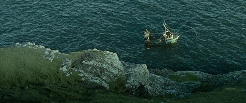 Ondine (2009) movie still: establishing — A rugged coastline features grassy cliffs meeting dark blue ocean water. A small fishing boat floats on…