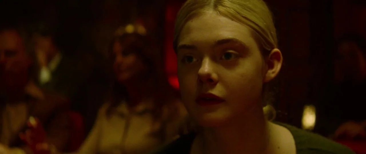 Low Down (2014) movie still: closeup — A dimly lit room with out-of-focus figures in the background suggests a social setting. A young woman with…