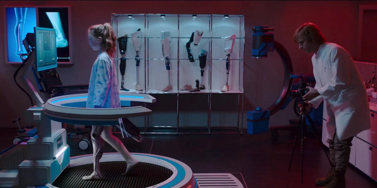 Mute (2018) movie still: wide — A dark, clinical space features a glass display case with various mechanical prosthetic limbs. A young girl uses a…