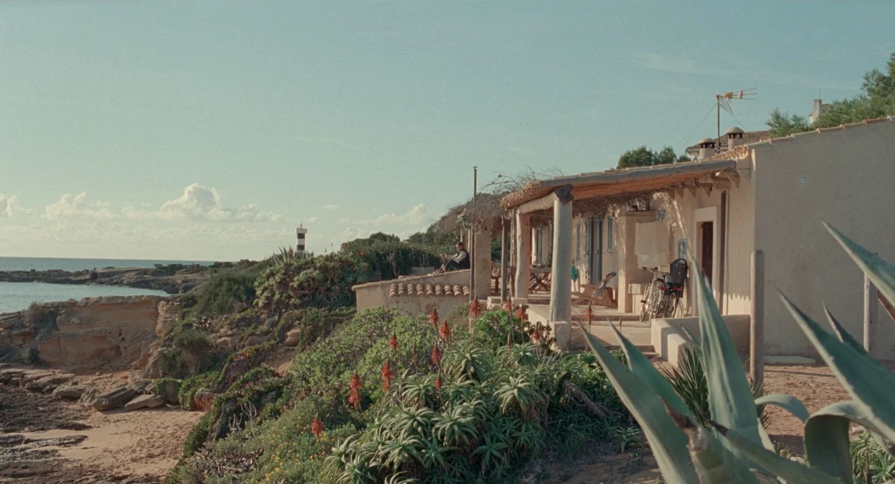 Non-Fiction (2018) movie still: wide — A coastal house with a covered patio overlooks a rocky beach and the sea. A distant lighthouse stands on the…