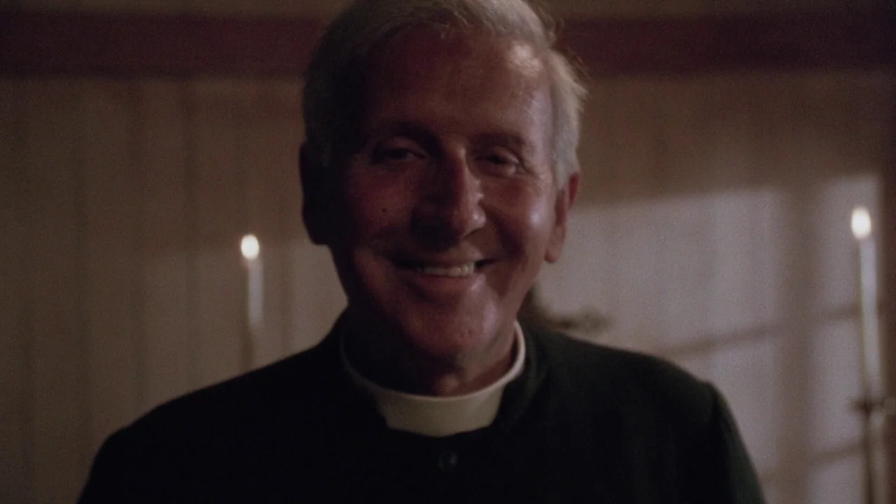 Insignificance (1985) movie still: closeup — A dimly lit room features wood paneling and two candles flanking the man. An elderly man in a clerical…