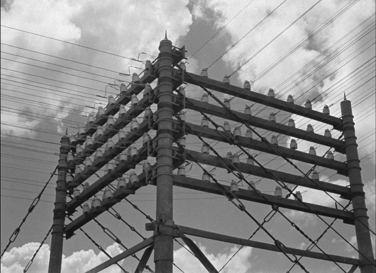 Pather Panchali (1955) movie still: wide — A complex metal utility pole stands against a cloudy, bright sky. Many ceramic insulators are attached to…