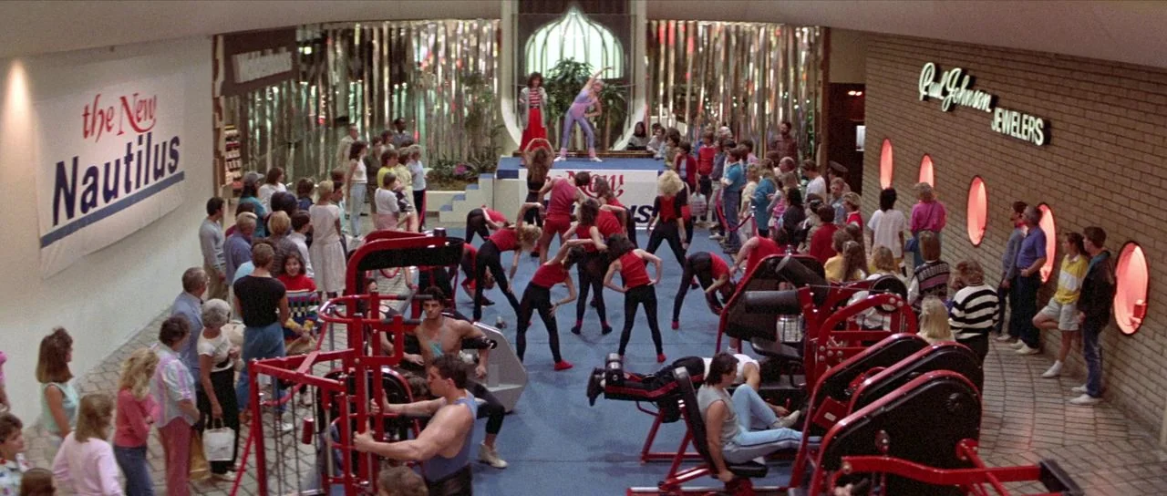 Bill and Ted’s Excellent Adventure (1989) movie still: wide — A crowded 1980s shopping mall features a large aerobic exercise event on display.…