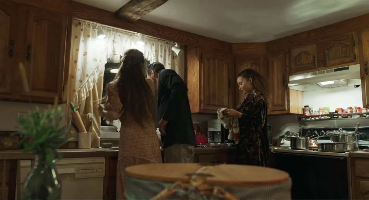 Salvation (2025) movie still: medium — A residential wood-cabinet kitchen features a large window with white curtains above the sink. Three people…