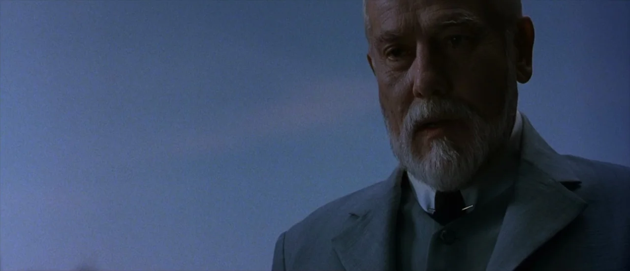 The Matrix Revolutions (2003) movie still: closeup — An elderly man in a formal grey suit stands outdoors at dusk. He looks down toward an area…