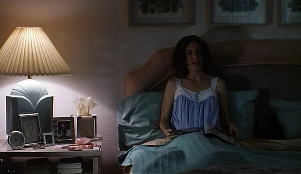 Safe movie still: medium — A bedroom at night featuring a woman sitting up in bed with a magazine. A table lamp with a patterned shade sits on a…