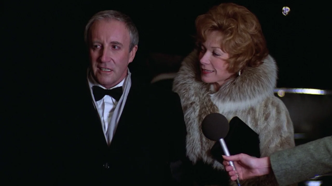 Being There (1979) movie still: medium — A dark night setting featuring an elderly man and woman in formal attire. A microphone is held in the…