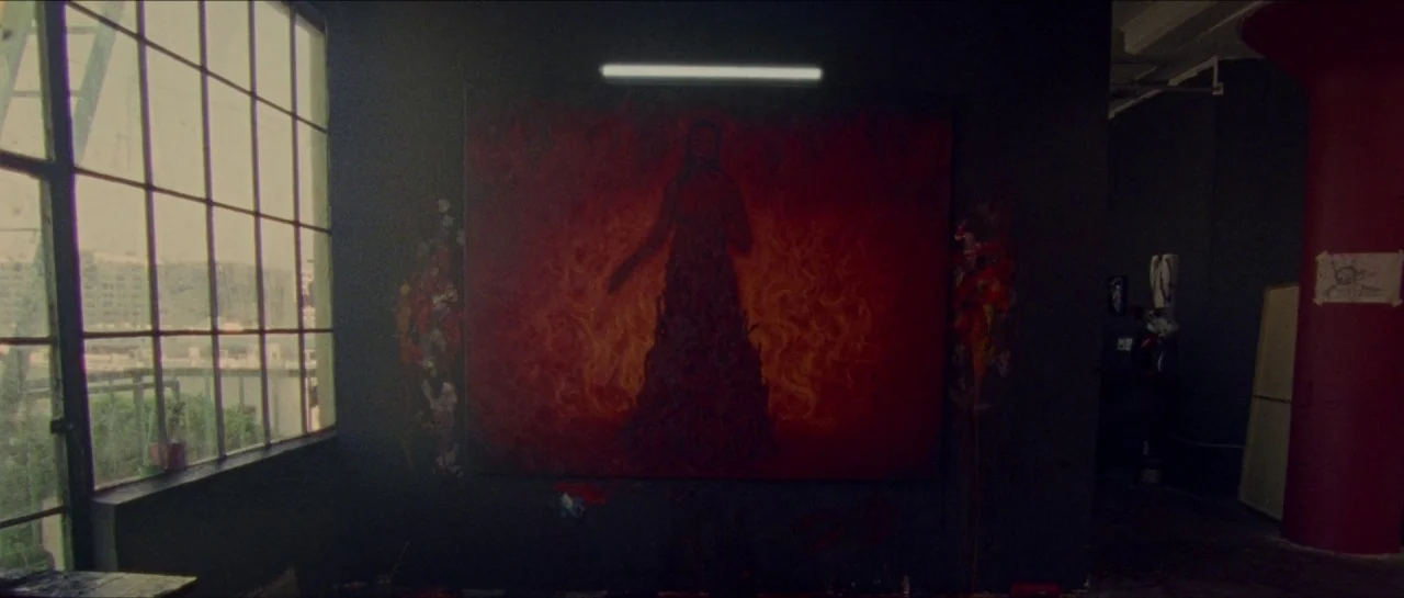 Bliss movie still: wide — A dark, industrial art studio features a large, vibrant red painting on the wall. The canvas depicts a dark figure amidst…