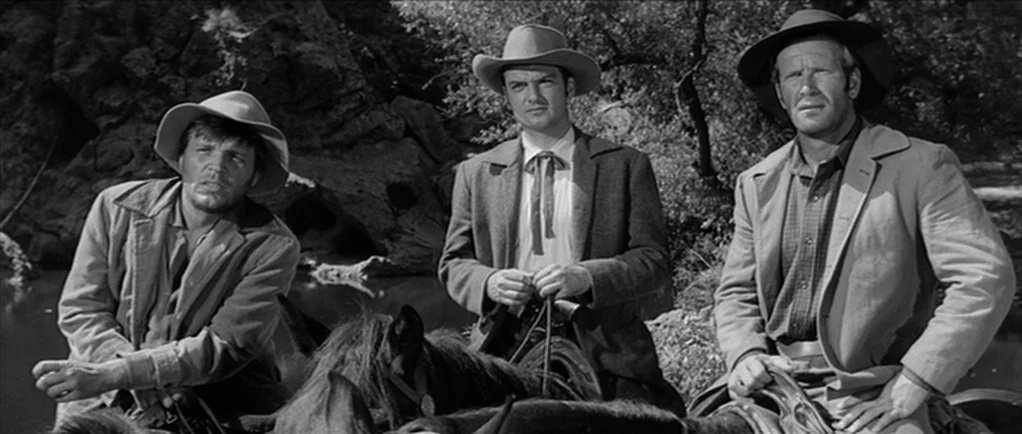 Love Me Tender movie still: medium — Three men ride horses along a riverbank surrounded by thick brush and trees. The men sit upright in their…