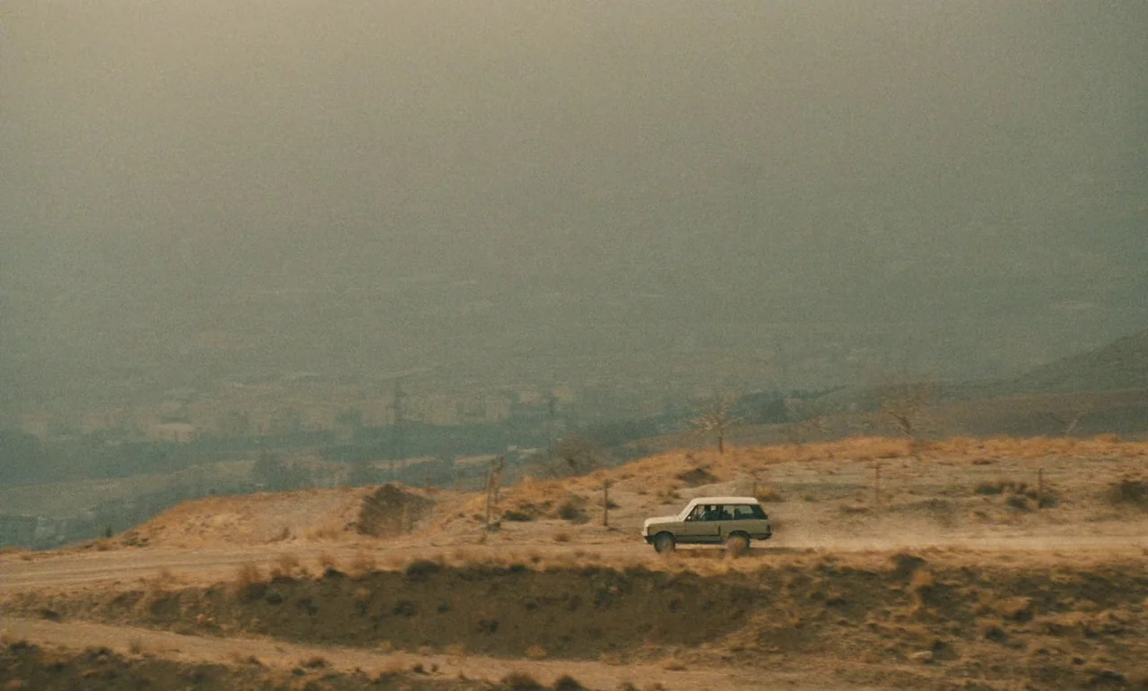 Taste of Cherry (1997) movie still: wide — A white vehicle drives across a dusty, dry landscape under a thick, hazy sky. The environment consists of…