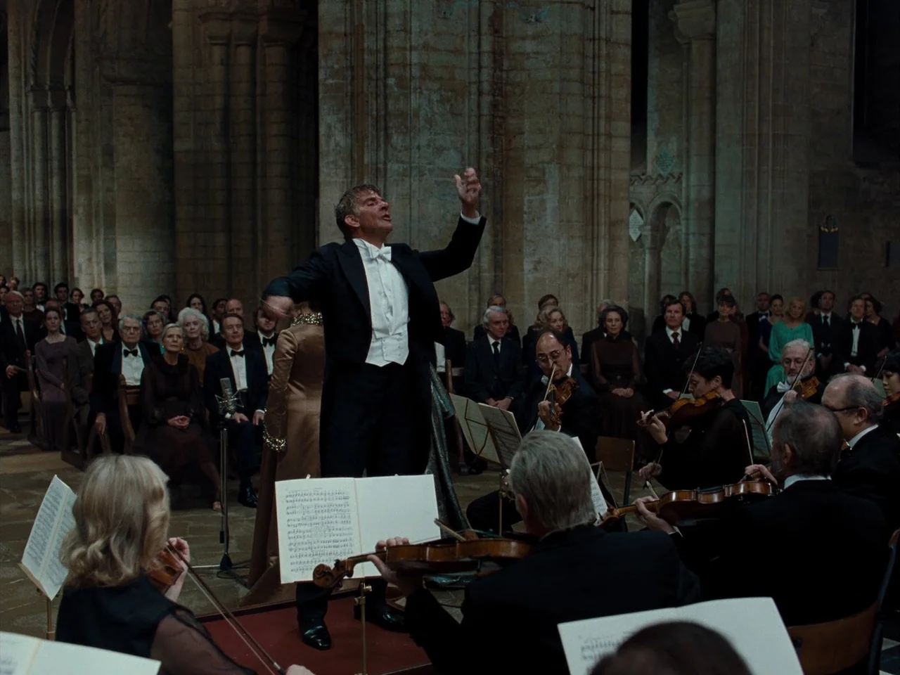 Maestro (2023) movie still: fullbody — A stone cathedral interior filled with a formal audience and an active orchestra. A male conductor wearing a…