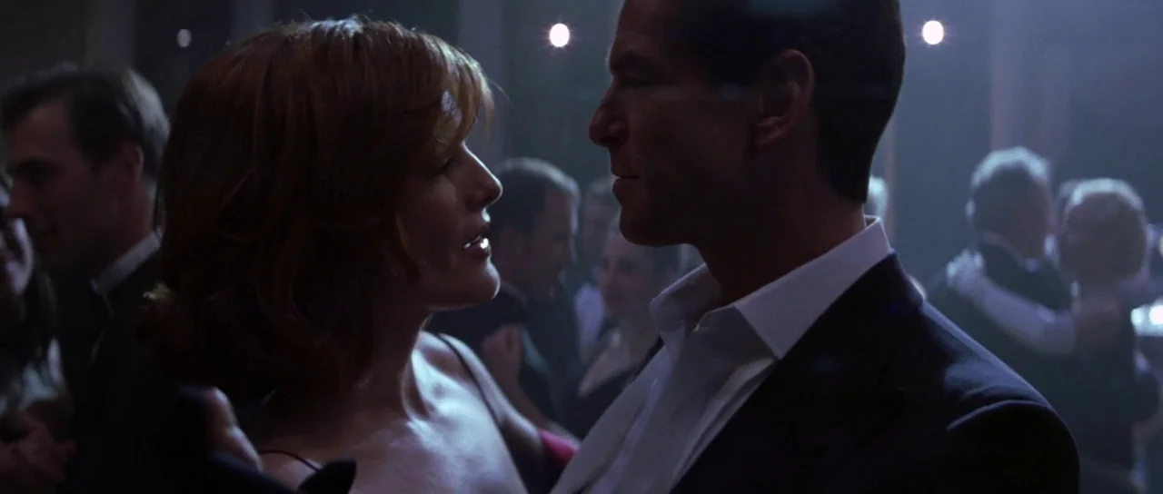 The Thomas Crown Affair (1999) movie still: medium — A dimly lit ballroom with figures dancing in the background. A man and a woman in formal attire…