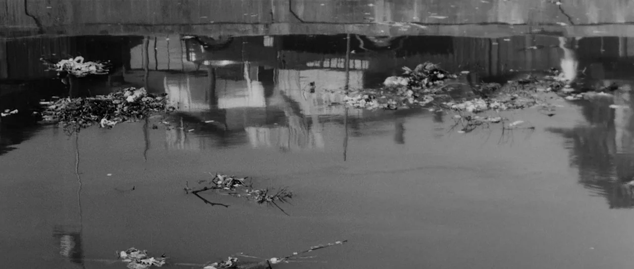 High and Low (1963) movie still: establishing — Stagnant canal water contains patches of floating waste, refuse, and submerged twigs. A concrete…