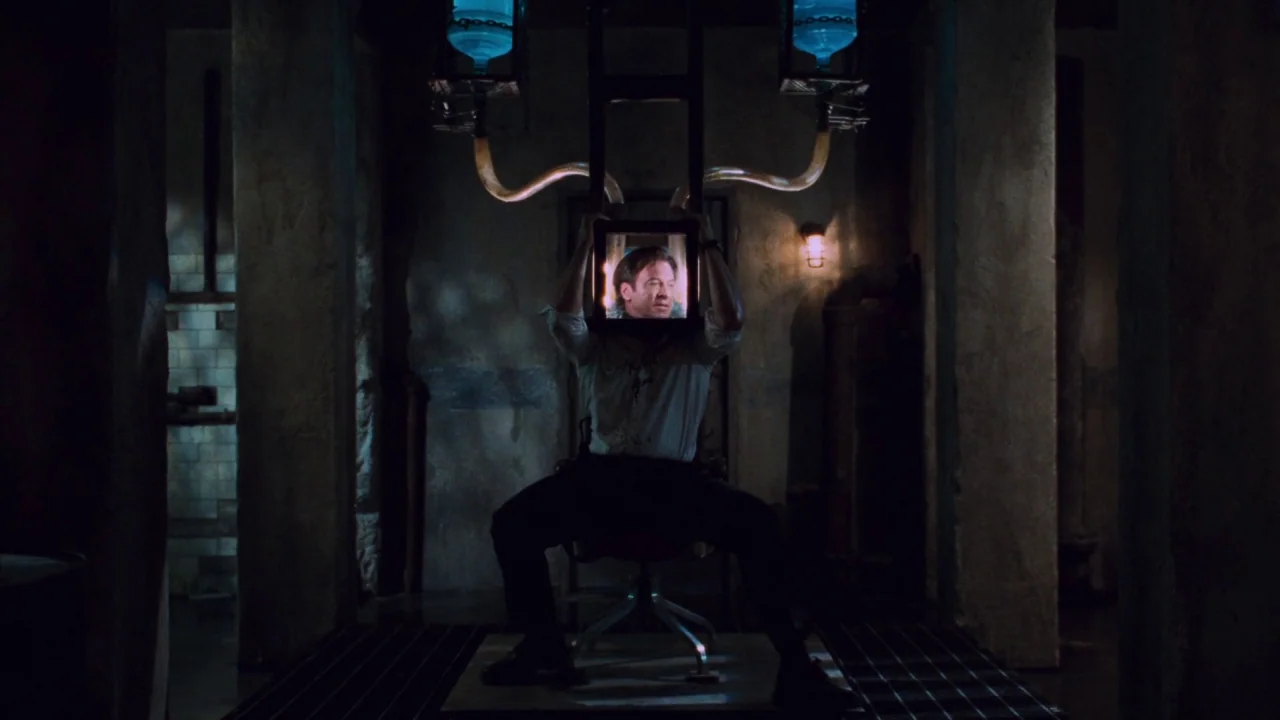 Saw V (2008) movie still: medium — An industrial space with concrete pillars and dark, cavernous surroundings. A man sits in a chair while holding a…