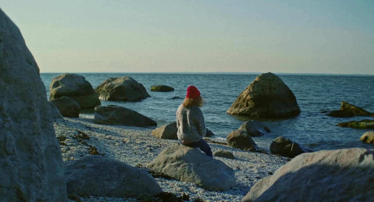 Blow The Man Down (2019) movie still: wide — A rocky beach with numerous large boulders along the shoreline meets the open water. A person wearing a…