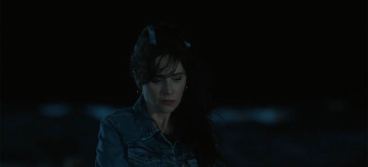 Merv (2025) movie still: closeup — A dark, nondescript outdoor location at night with no visible background details. A woman in a denim jacket gazes…
