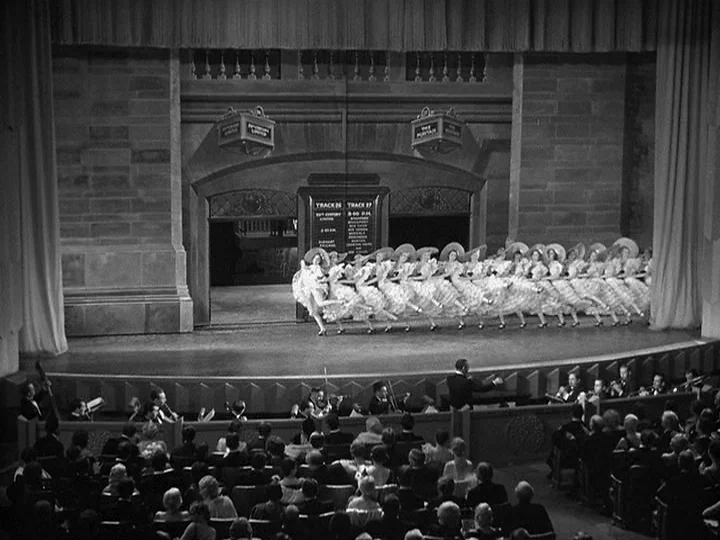 42nd Street (1933) movie still: wide — A theatrical stage depicts a railway station backdrop with a line of dancing women. Dancers in voluminous…