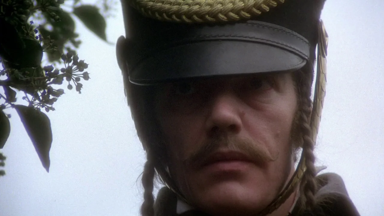 The Duellists (1977) movie still: closeup — An outdoor meadow with foliage visible in the periphery under an overcast sky. A man in a…