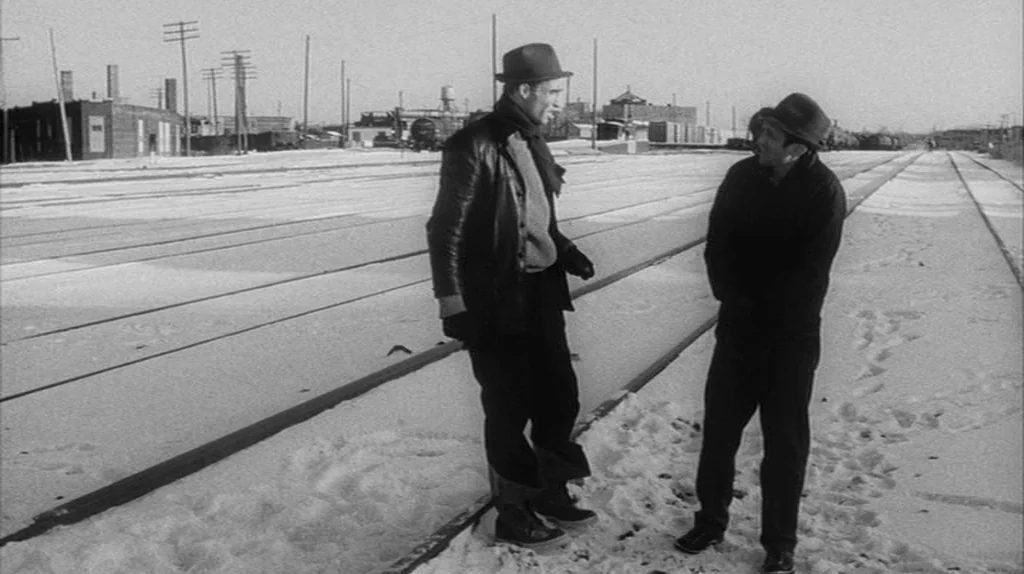 Stranger Than Paradise (1984) movie still: fullbody — A snowy train yard features long parallel tracks extending toward industrial brick buildings.…