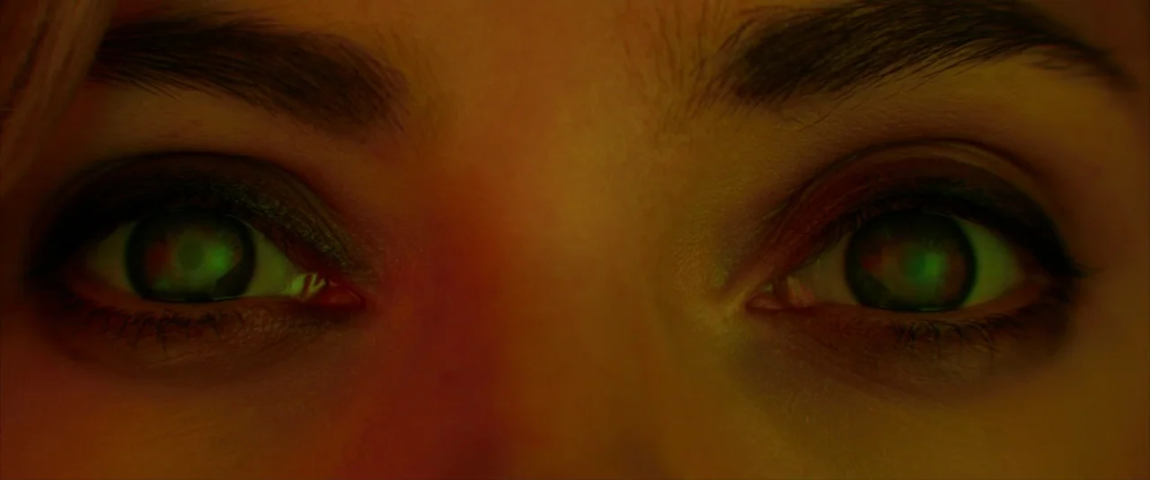 Lucy (2014) movie still: closeup — A tight extreme closeup of a human face focused entirely on the eyes. The dark pupils reflect a bright green…