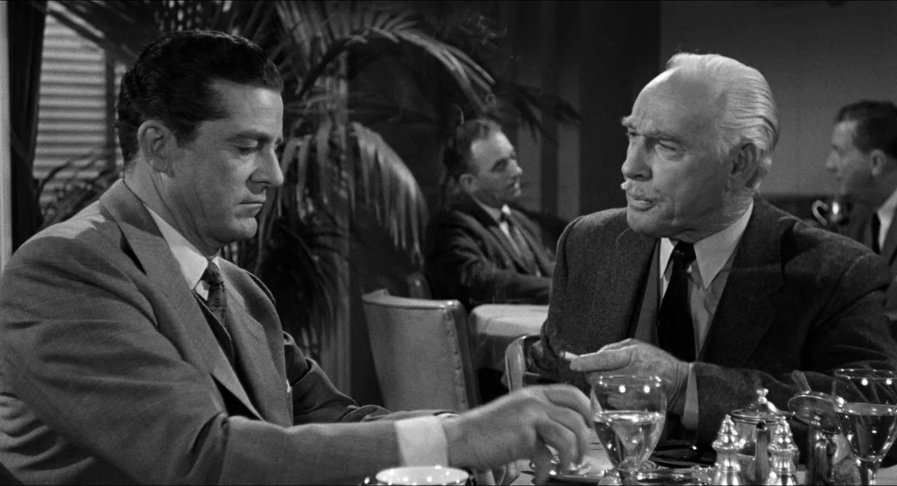 The Fearmakers (1958) movie still: medium — A mid-century restaurant dining room with a potted plant in the background. Two men in suits sit at a…