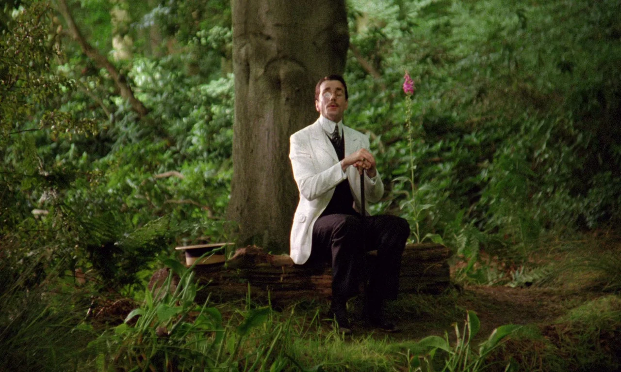 A Room With A View (1985) movie still: fullbody — A man sits on a fallen log within a dense green thicket of ferns. The man rests both hands on a…