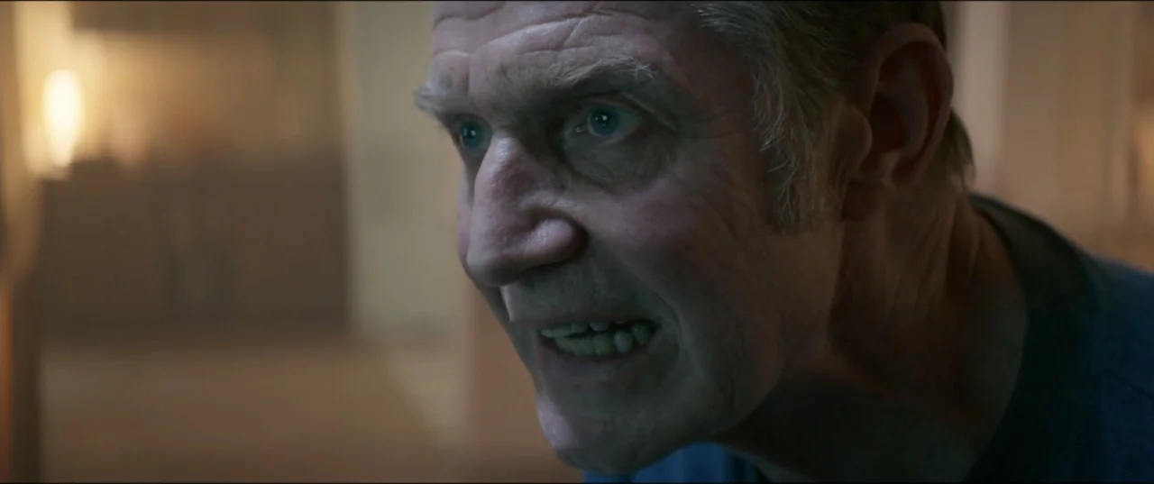 Utopia (2013) movie still: closeup — An interior space is dimly lit with blurred background elements suggesting a domestic setting. An elderly man…
