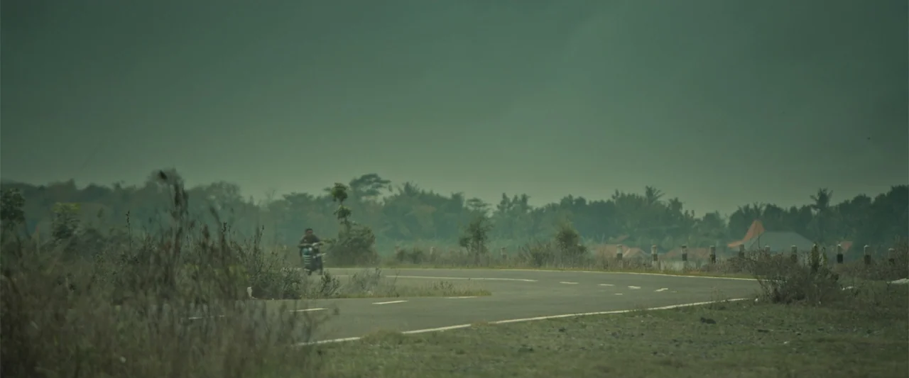 The Unholy (2022) movie still: wide — An asphalt road curves through a flat, rural landscape under a heavy overcast sky. A lone motorcyclist rides…