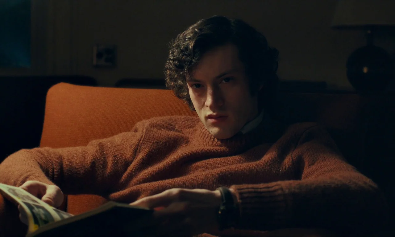 The Holdovers (2023) movie still: medium — A dark, dimly lit room features a person sitting in a burnt-orange fabric armchair. The young man with…