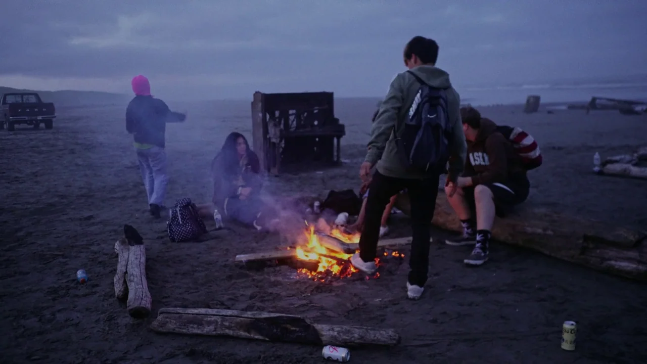 Gasoline Rainbow (2023) movie still: wide — A group of teenagers gathers around a small campfire on a misty, desolate beach. An upright piano sits…