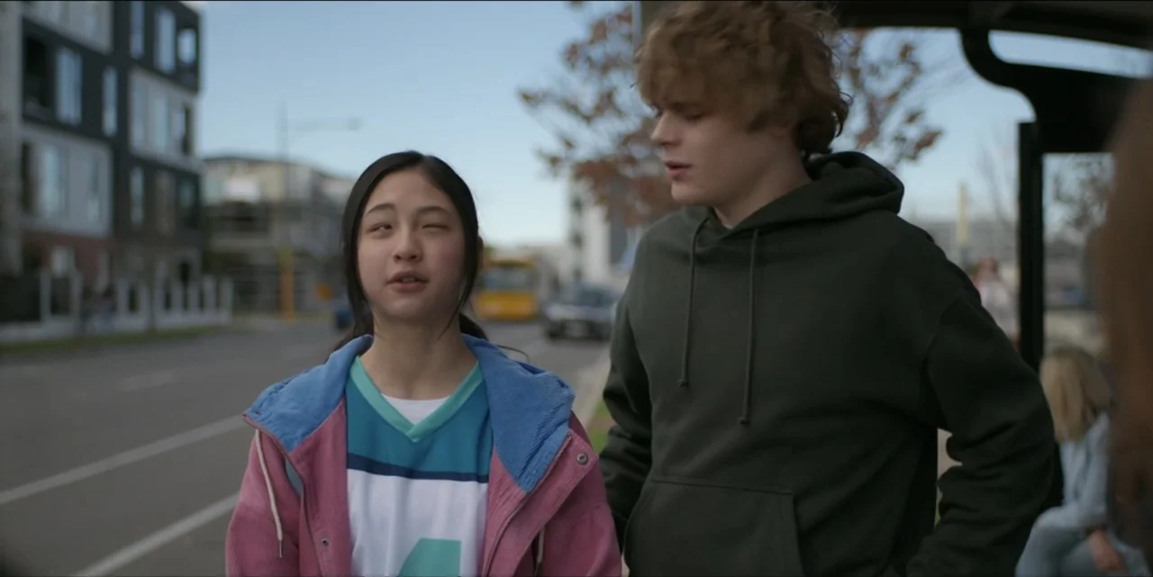 Bring Her Back (2025) movie still: medium — An urban sidewalk with a bus shelter and modern apartment buildings in the background. A teenage girl…