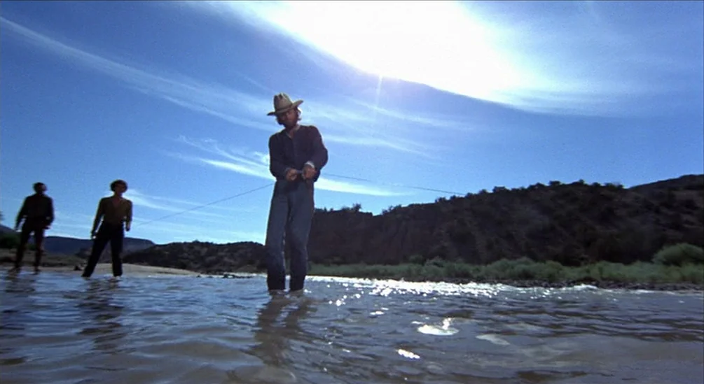 The Hired Hand (1971) movie still: fullbody — A wide, shallow river flows through an arid, rocky desert landscape under a clear sky. A man stands in…
