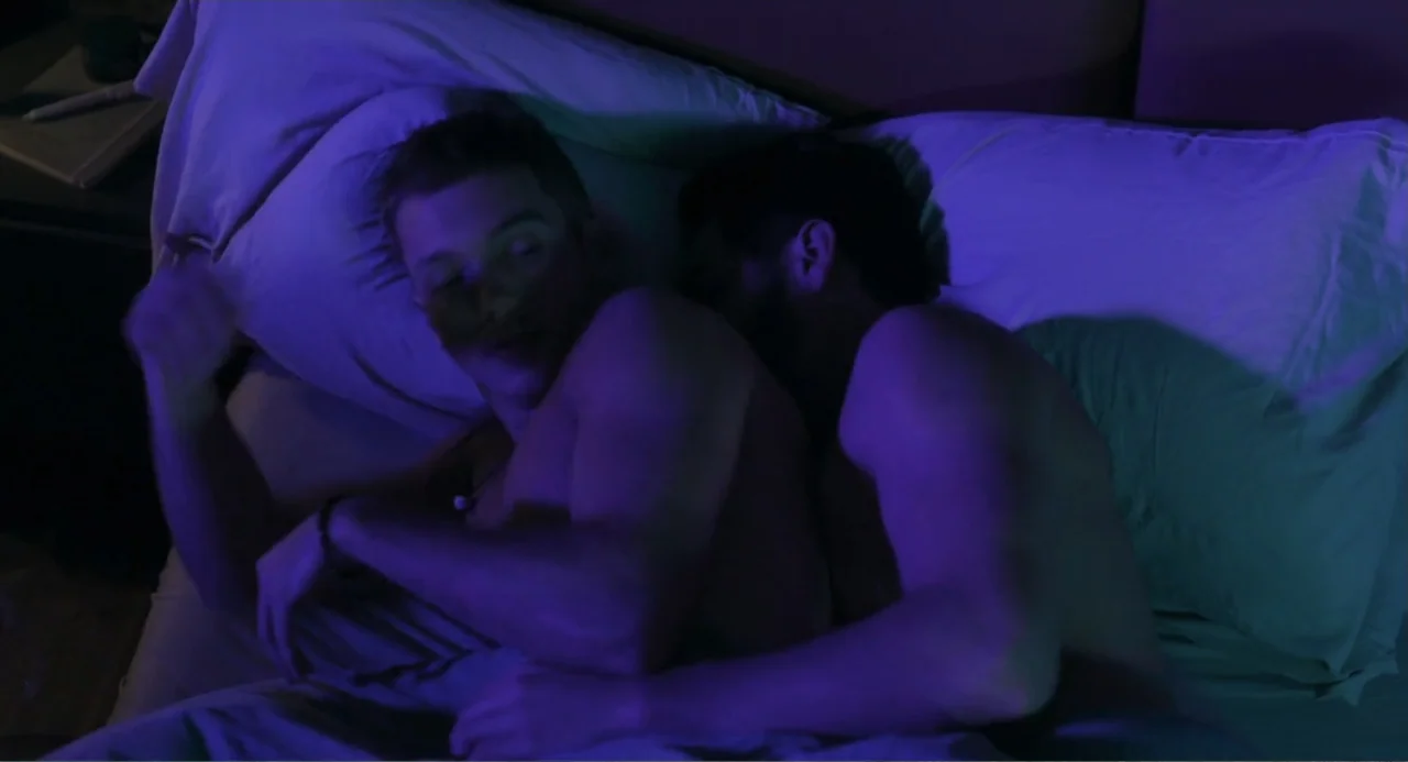 Straight (2024) movie still: medium — A dark bedroom interior features two men lying together under white bedding sheets. The men rest against…