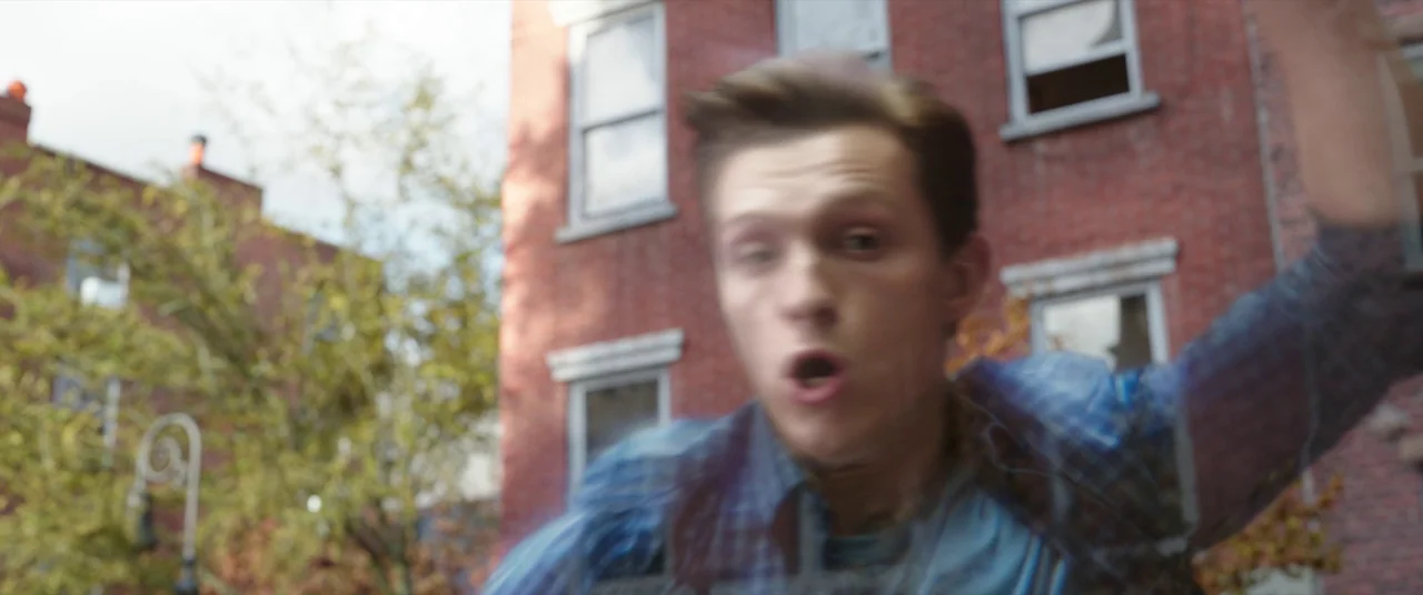 Spider-Man: No Way Home (2021) movie still: closeup — A residential street features a red brick building with multiple white-framed windows. A young…