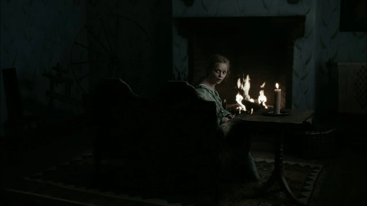 Sleepy Hollow (1999) movie still: medium — A dimly lit room with blue patterned wallpaper and a large spinning wheel in shadow. A young woman sits…