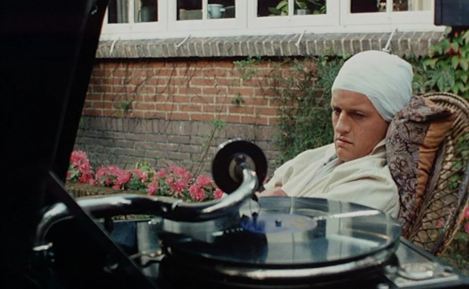 Soldier of Orange (1977) movie still: medium — A man wearing a head bandage sits in a wicker chair outdoors. A foreground record player plays music…