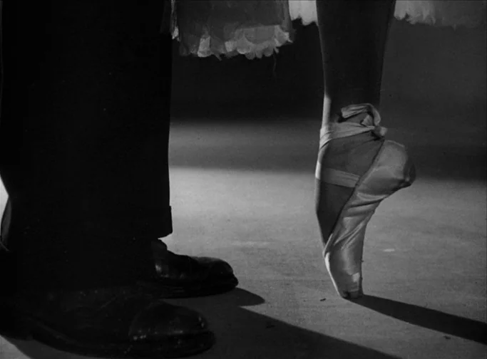 Summer Interlude (1951) movie still: closeup — A dark stage floor shows the feet of a man and a dancing ballerina. The ballerina rests on one pointe…
