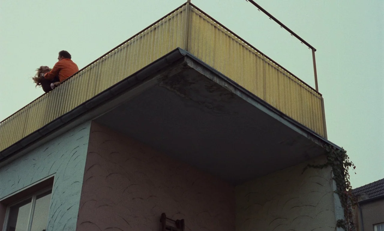 Wrong Move (1975) movie still: wide — A modern building exterior features a yellow balcony railing under a flat grey sky. A man in an orange jacket…
