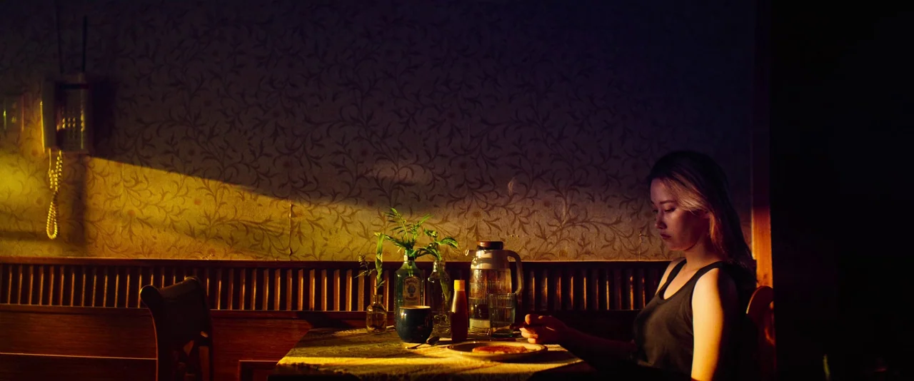 Ballerina (2023) movie still: medium — An dimly lit diner booth features vintage floral wallpaper and dark wood accents. A woman sits alone at a…