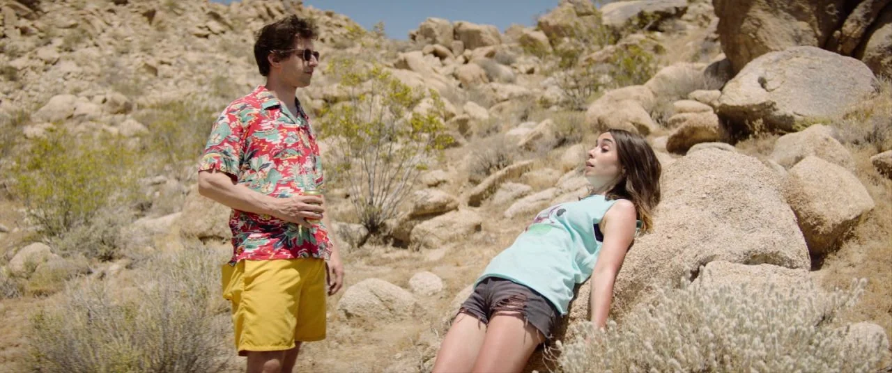 Palm Springs (2020) movie still: fullbody — A rugged desert landscape features large tan boulders and sparse brush under bright sun. A man stands…