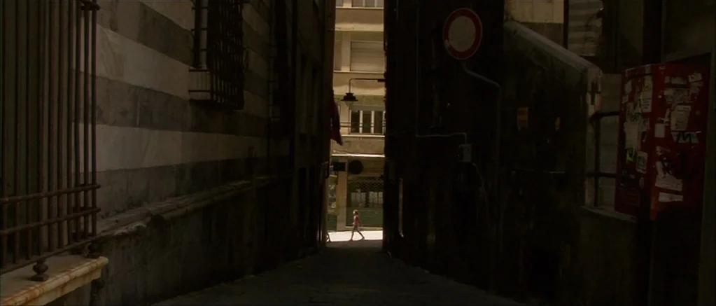 Genova (2008) movie still: wide — A narrow, dark urban passageway with high stone walls and a distant street opening. A single person walks across…