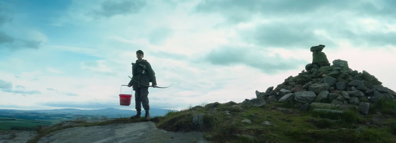 28 Years Later (2025) movie still: fullbody — A grassy ridge under an overcast sky features a stone cairn on the right. A young boy stands on the…