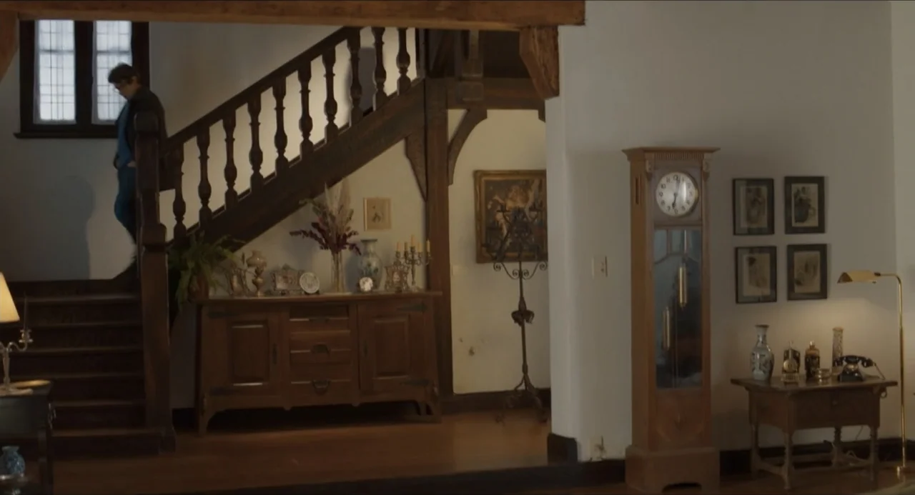 Echoes Of A Crime (2025) movie still: wide — A traditional wood-paneled foyer features a staircase, a grandfather clock, and antique sideboards. A…