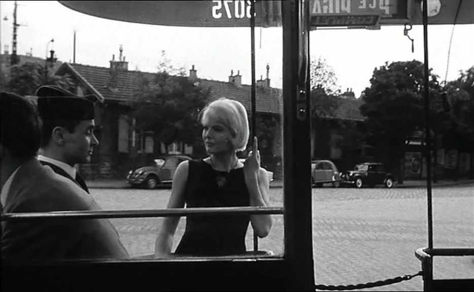 Cléo from 5 to 7 movie still: medium — The interior of a bus looking out onto a cobblestone street with small cars. A woman in a black dress talks…