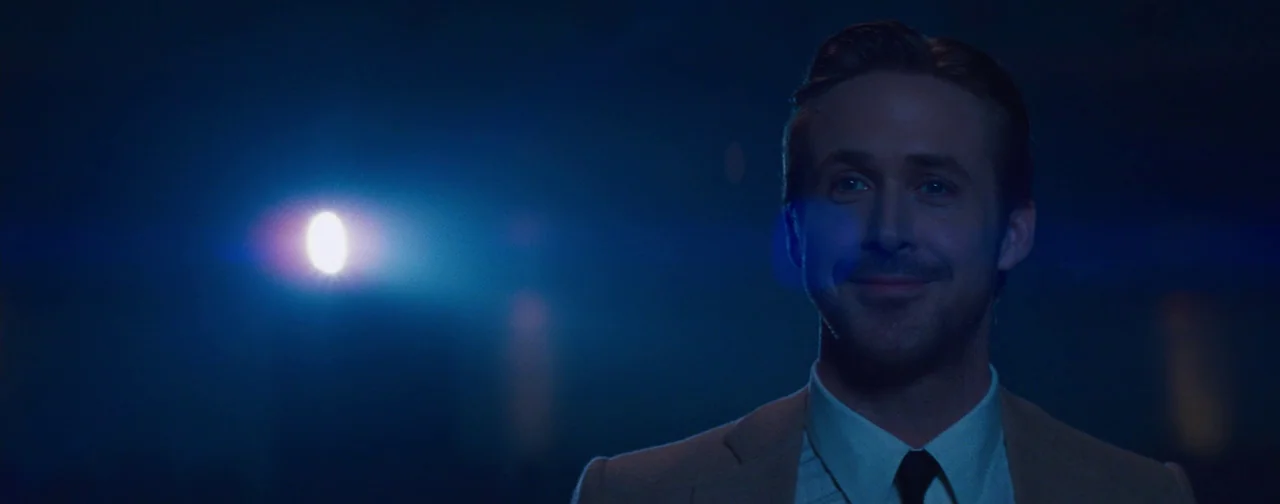 La La Land (2016) movie still: medium — A dimly lit room is illuminated by a single glowing point of light. A man in a suit stands in the foreground…