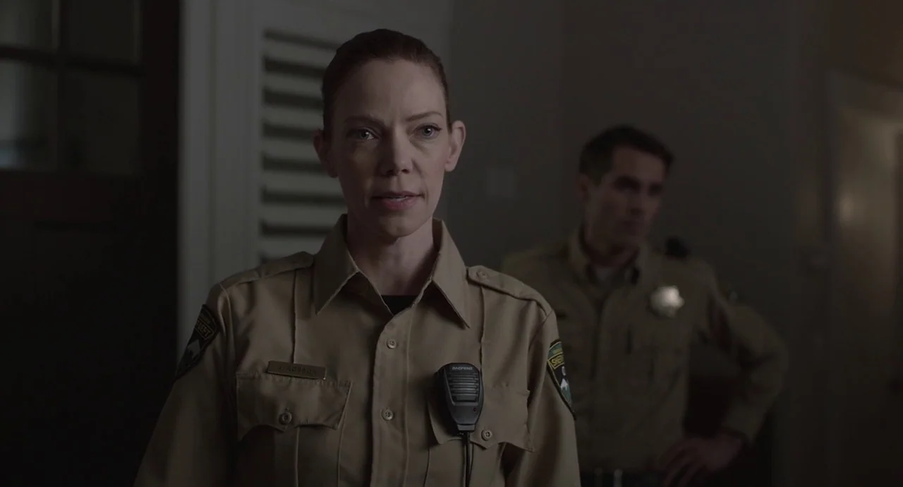 The Wolf of Snow Hollow (2020) movie still: medium — Two law enforcement officers stand inside a dim, sparsely decorated administrative room. The…