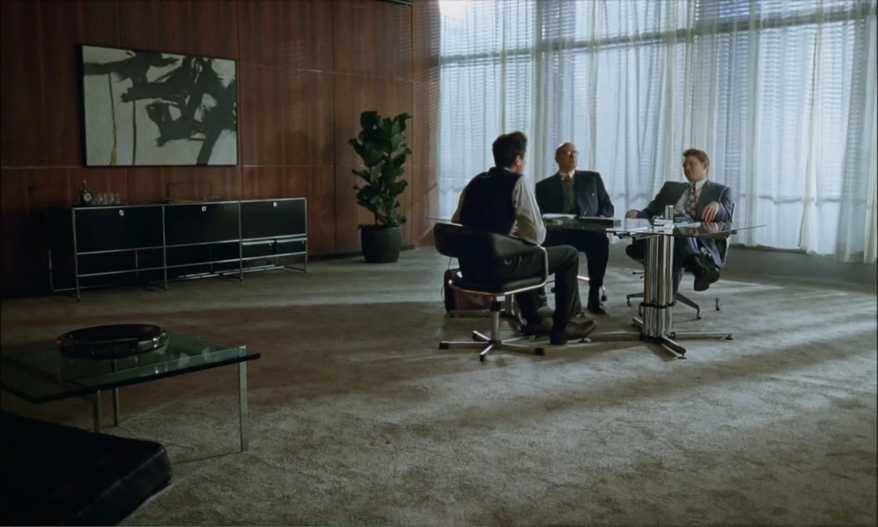 The Brutalist (2024) movie still: wide — A spacious office features wood paneling and large windows with floor to ceiling blinds. Three men sit…