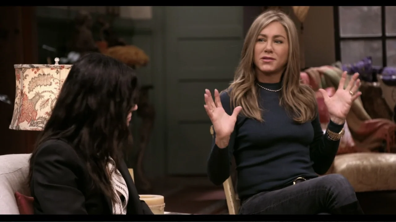 Friends: The Reunion (2021) movie still: medium — A cozy, well-lit domestic living room contains fabric armchairs and a decorative table lamp. A…