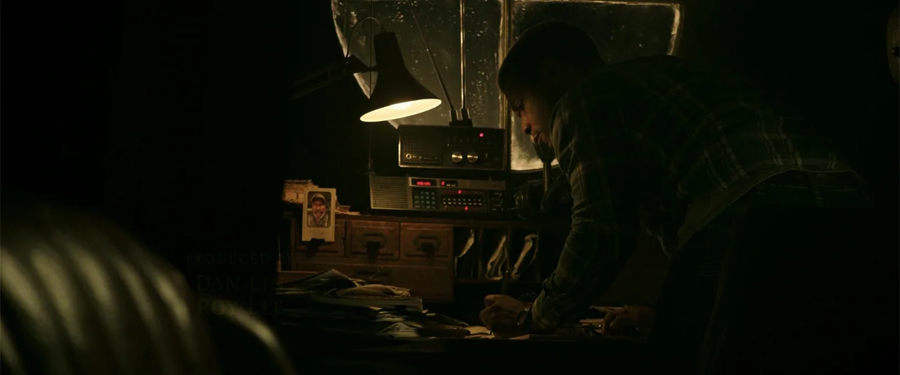 K.G.F: Chapter 2 (2022) movie still: medium — A dark office contains a wooden desk covered in papers and a vintage radio. A man bends over the desk…